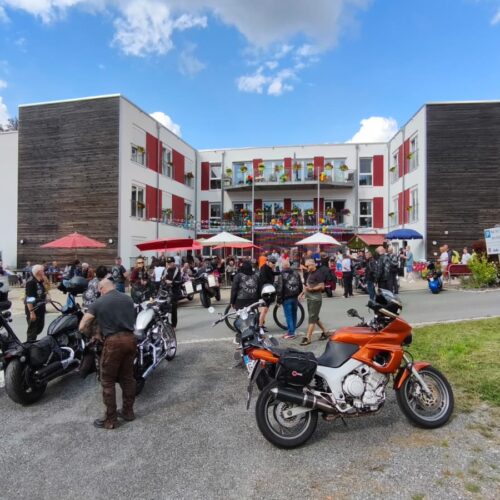 Mehrere Motorradfahrer stehen lachend vor einem modernen, hellen Gebäude mit roten Akzenten