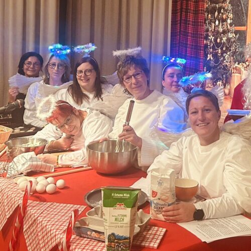 Eine fröhliche Gruppe Frauen in Engelskostümen backt mit Weihnachtsmann am roten Tisch