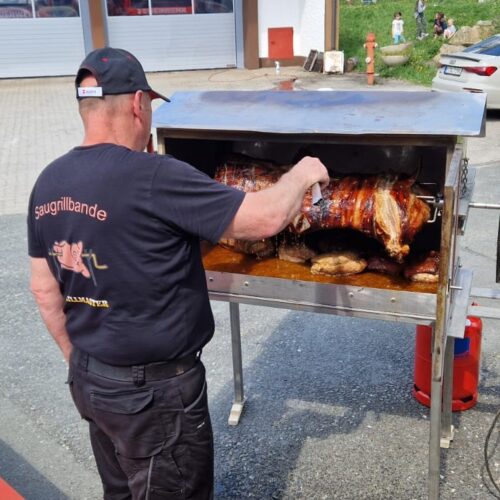 Ein Mann mit schwarzem Outfit wendet ein gegrilltes Spanferkel am Grillgerät