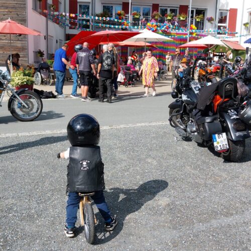 Ein Kind mit schwarzer Weste und Helm fährt Laufrad zu einer Gruppe von Bikern