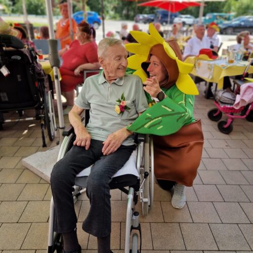 Ein älterer Mann im Rollstuhl lächelt neben einer Person im Sonnenblumenkostüm in Grün und Gelb