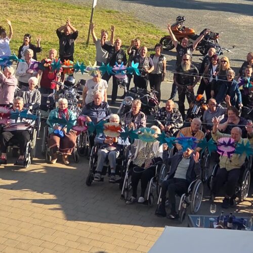 Eine Gruppe älterer Menschen im Rollstuhl und ihre Betreuer winken fröhlich im Sonnenschein