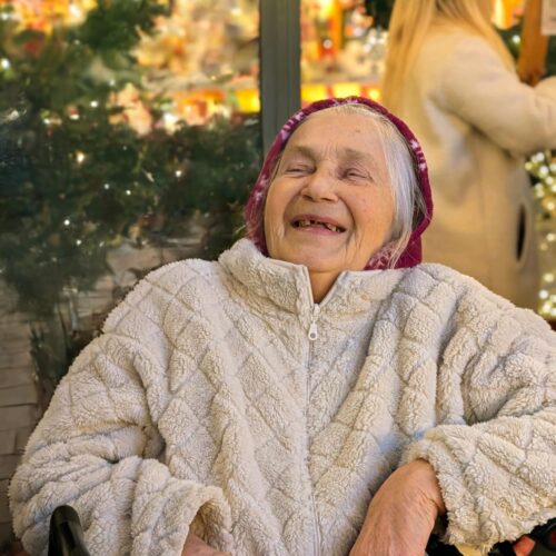 Ältere Frau mit weißem Haar und fröhlichem Lächeln sitzt in heller Jacke vor Lichtern