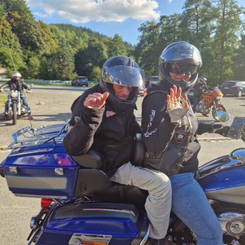 Zwei Personen auf blauem Motorrad lächeln freundlich und winken Richtung Kamera