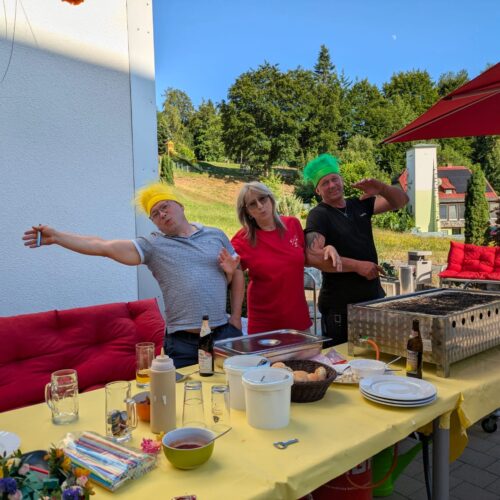 Drei Personen mit bunten Perücken posieren lachend neben einem Grill auf einer Terrasse