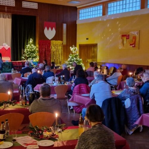 Mehrere Personen sitzen in einem festlich geschmückten Saal mit leuchtenden Tannenbäumen und Kerzen, während eine kleine Gruppe auf der Bühne spricht