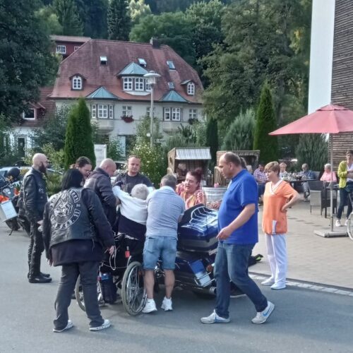 Mehrere Menschen lachen draußen, eine Person im Rollstuhl wird von Motorradfahrern umgeben