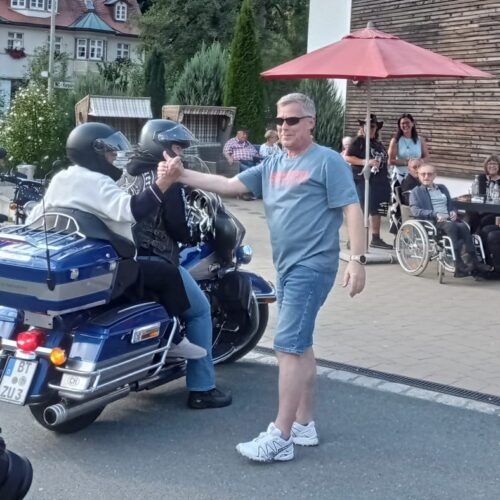 Ein Mann in grauem T Shirt und Shorts gibt einer Motorradfahrerin mit dunklem Helm die Hand