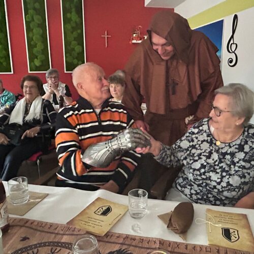 Ein älterer Mann mit Ritterhandschuh lacht freundlich neben einer Dame, ein Mönch steht dahinter