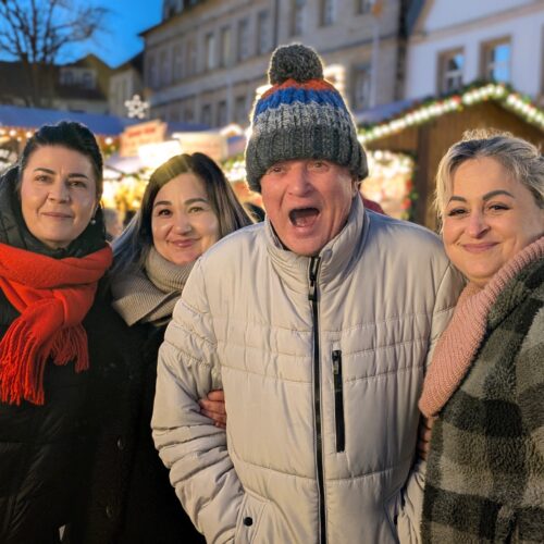 Vier Erwachsene stehen fröhlich zusammen im Wintermantel auf einem Weihnachtsmarkt