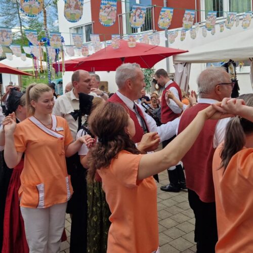Eine Gruppe fröhlicher Menschen tanzt im Freien, einige in Tracht, andere in orangefarbener Kleidung