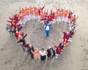 Menschen verschiedener Altersgruppen stehen in Herzform und lächeln freundlich hoch