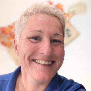 Eine lächelnde Person mit kurzem, blondem Haar und blauer Kleidung vor Herbstdeko