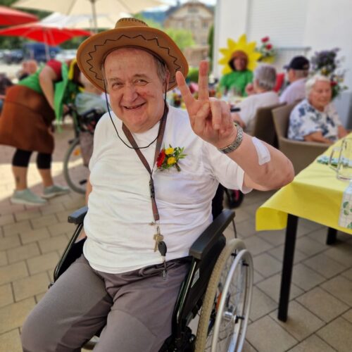 Ein älterer Mann im Rollstuhl lächelt freundlich und zeigt das Victory Zeichen, trägt einen braunen Cowboyhut und ein weißes TShirt mit einer gelben Blume an der Brust, im Hintergrund sitzen mehrere Menschen unter roten Sonnenschirmen bei einer sommerlichen Feier