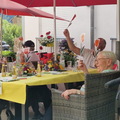 Eine ältere Frau mit Brille lächelt freundlich an einem gelb gedeckten Tisch unter roten Schirmen