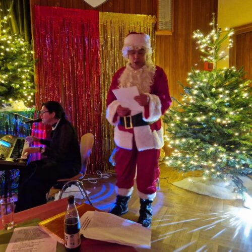 Ein Mann im Weihnachtsmannkostüm liest mit ernster Miene neben blinkenden Bäumen vor