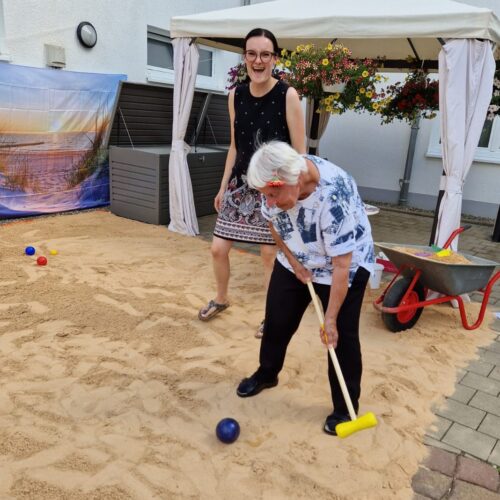 Eine ältere Frau mit weißem Haar spielt lachend Boccia im Sand vor einer lachenden Frau