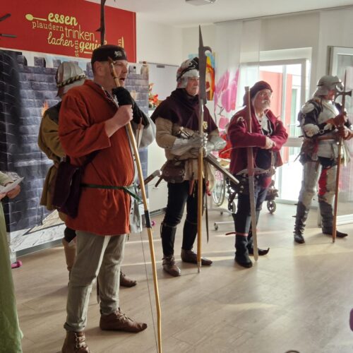 Sieben Menschen in mittelalterlicher Kleidung und mit Bögen stehen lächelnd im hellen Raum