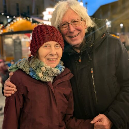 Zwei ältere Menschen lächeln herzlich nebeneinander auf einem festlich beleuchteten Weihnachtsmarkt