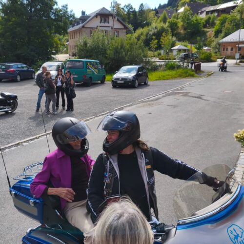 Zwei Personen mit Helmen auf einem blauen Motorrad lächeln sich in einer Dorfstraße an