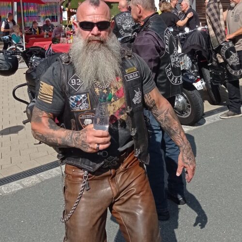 Ein bärtiger Mann mit Sonnenbrille und Lederkleidung steht lächelnd auf einem Bikertreffen